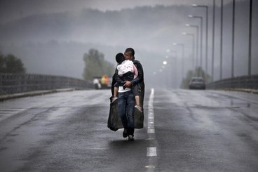 Foto di Yannis Behrakis, con padre e figlio migranti bagnati dalla pioggia. Il padre porta in braccio il figlio e insieme camminano su una strada (o un ponte)