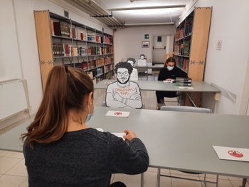 Sala di lettura della Biblioteca di Scienze dell'Educazione con studenti seduti ai tavoli e sagome raffiguranti Patrick Zaky accanto a loro