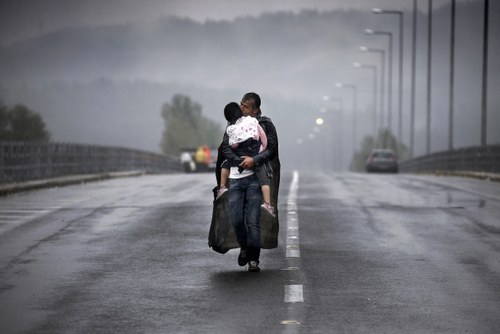 Immagine simbolo del convegno e della mostra: foto di Yannis Behrakis, con padre e figlio migranti bagnati dalla pioggia. Il padre porta in braccio il figlio e insieme camminano su una strada (o un ponte)