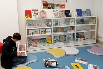 LAboratorio di Letteratura per l'Infanzia: vetrina espositiva con albi illustrati all'interno e sopra, tappeto e cuscini a disposizione per la lettura e consultazione e utente del Laboratorio immerso nella lettura